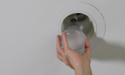 A person installing a recessed light bulb in a fixture on the ceiling.