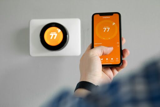 Homeowner's hand using phone to adjust the temperature of the smart thermostat in the background