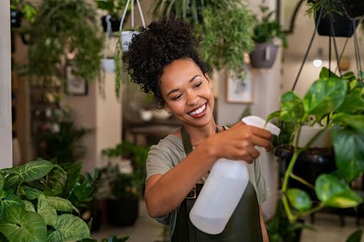 A smiling woman spraying water on a hanging houseplant.